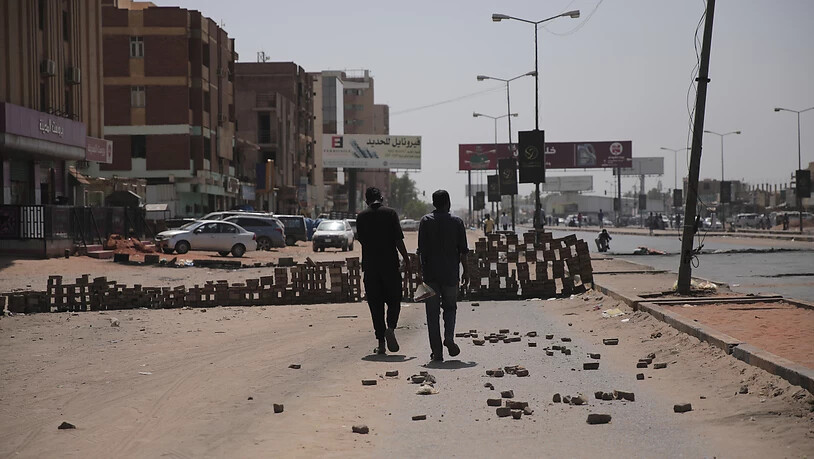 Zwei Personen gehen auf einer Straße auf der Steine liegen und eine behelfsmäßigen Barrikade aufgebaut ist. Nach dem Militärputsch am 25. Oktober 2021, bei dem das Militär die Macht über das Land übernommen hat, ist der entmachtete Ministerpräsident…