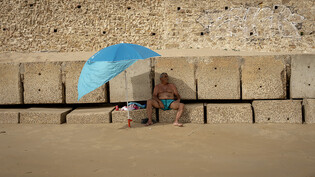 Ein Mann sitzt in Cadiz auf einem Wellenbrecher und schützt sich vor der Sonne unter seinem Sonnenschirm an einem Strand. Foto: Emilio Morenatti/AP/dpa